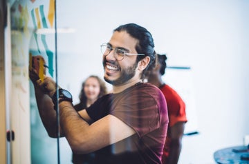 Side view of cheerful bearded man in glasses writing memos on sticky notes while creating plan on glass board in office with colleagues