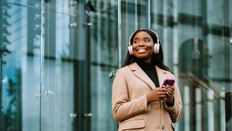 Young happy black businesswoman using cellphone and headphones while walking outdoors