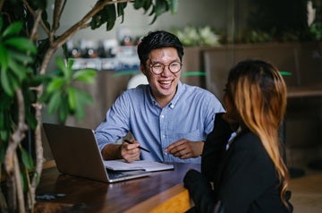 A confident and handsome young man is interviewing a female candidate for a job. They are sitting together in a table at a trendy coworking space. He is laughing and having a relaxed conversation.